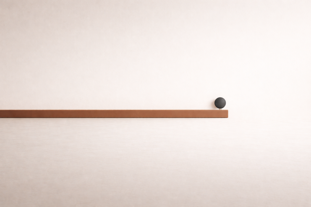 Minimalist still life featuring a horizontal clay-brown beam and a small charcoal dot above it on a warm sand surface, symbolizing emotional restraint and pause before reaction.